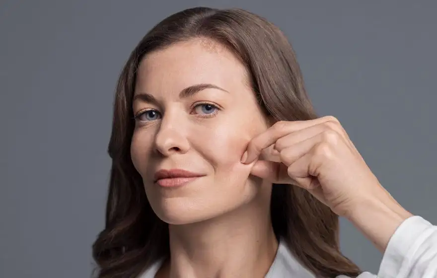 Uma mulher com pele visivelmente saudável e luminosa, de cabelos castanhos claros, pinça delicadamente a bochecha com os dedos, sorrindo levemente. A ação destaca a elasticidade e a firmeza da pele, ilustrando os benefícios do bioestimulador de colágeno na melhora da textura, estimulando a produção natural de colágeno e elastina.