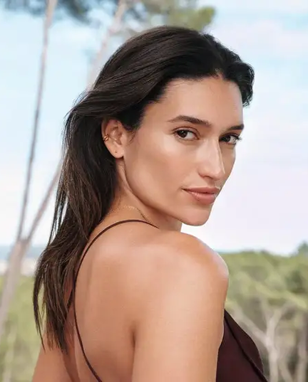 Uma mulher morena de cabelos lisos, usando uma blusa de alças finas marrom. Ao fundo, uma área verde desfocada
