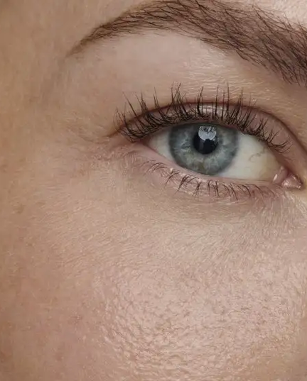 Uma imagem em close-up do rosto de uma pessoa, focando no olho esquerdo. A pele tem um tom claro e é visível a textura com alguns poros e pequenas sardas ou manchas claras. O olho, de íris azul-acinzentada, está aberto e centralizado na imagem, com a pupila escura refletindo uma fonte de luz. Os cílios superiores são longos e escuros, com um leve volume, e os cílios inferiores são mais curtos. A sobrancelha é bem definida e de cor castanha. A área da bochecha e parte da orelha são visíveis no lado esquerdo`