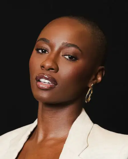 Uma mulher de pele escura e cabelo raspado, com um corte de cabelo bem curto, posa de frente para a câmera contra um fundo preto. Ela está usando um blazer de cor clara, possivelmente bege ou creme, e um brinco dourado em formato de onda na orelha direita. Seus lábios, que parecem ter sido realçados por preenchimento labial, estão levemente abertos, revelando os dentes, e têm um brilho labial que reflete a luz. A maquiagem realça seus olhos e maçãs do rosto, dando-lhe uma aparência elegante e confiante.