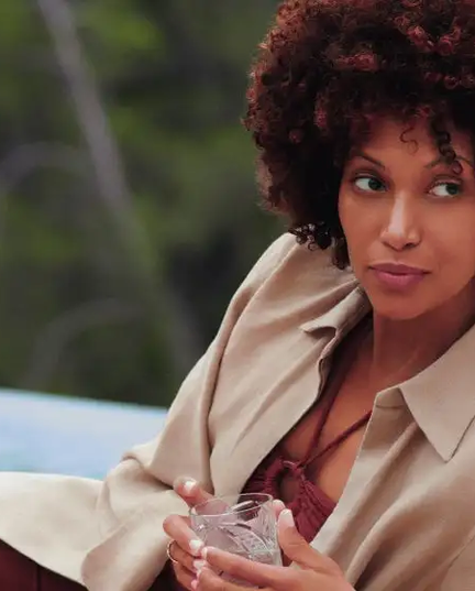 Uma mulher negra de cabelos volumosos e cacheados, usando uma camisa bege e calça vinho, relaxa em um sofá claro ao ar livre perto de uma piscina, com uma bebida em um copo transparente nas mãos. Ela olha para o lado, com uma expressão serena e confiante, resultado do uso do bioestimulador de colágeno Sculptra. Ao fundo, uma área verde desfocada